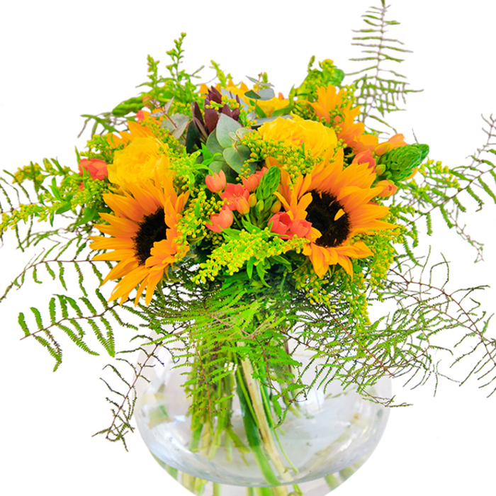 At first glance this bouquet reads as a sunlit chorus: vibrant sunflowers and yellow garden roses arranged with a natural economy that still feels rich and abundant. The sunflowers, with their thick, velvety petals graduating from golden yellow to orange, frame somber brown centres that give depth and structure. Intermingled, garden roses open in compact rosettes, their plush textures catching light and creating soft highlights. Small clusters of red Hypericum berries gleam amid the foliage, their glossy roundness providing lively contrast against the matte petals. The greenery is a layered study in texture-delicate, lace-like ferns spill gently downward; sprigs of light green filler, likely solidago, add tiny clustered blooms that whisper of late summer meadows; and upright green spikes give a subtle architectural lift. Cool eucalyptus leaves in a muted blue-green hue peek through, offering both tonal relief and a faint herbal scent that tempers the bouquet's sweetness. The round, clear glass vase, reminiscent of a fishbowl, displays the stems submerged in clean water, the overlapping lines of stalks visible as a compositional element of their own. This arrangement, ideal for a central table or a sunny windowsill, evokes the breezy, open feeling of a walk through Bayswater's Porchester Square on a warm afternoon. Crafted with attention to proportion and scent, it reads as a bright, optimistic gift for anniversaries, celebrations, or simply to send a slice of summer to a friend.