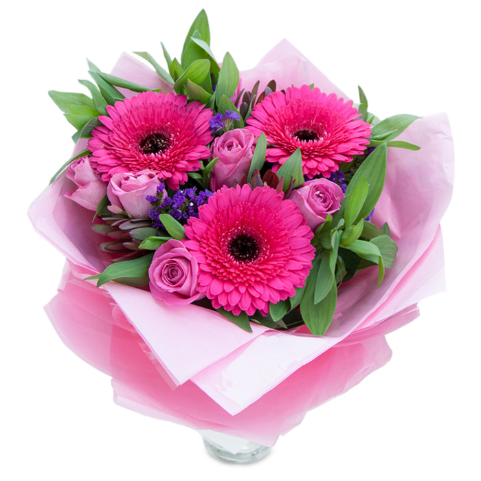 There is a playful elegance here: three wide-eyed Gerbera daisies in a confident fuchsia hue burst forward, their numerous petals arranged in neat concentric rows around dark centres that add drama and definition. Curling around them, dusty-rose to lavender-pink roses open with layered, velvety folds that soften the palette and invite a closer breath. Floating between the main flowers, delicate sprays of purple statice create airy ribbons of texture, like a small field of wildflowers tucked into the design. The composition is anchored by lush green foliage - glossy, substantial leaves that recall salal or eucalyptus - giving the bouquet a fresh, slightly herbal scent and a sense of botanical balance. The blooms rest in a clear glass vessel whose top rim is visible beneath an inner wrap of pale pink matte tissue and an outer sheet of sheer fuchsia-pink cellophane; the cellophane's subtle shimmer and crinkles catch the light of a soft morning and highlight the arrangement's pink tones. I imagine this placed on a kitchen island near Westbourne Grove or the edge of Hyde Park, where it would lift the room with colour and fragrance. Crafted with attention to form and freshness, this bouquet reads as both intimate and celebratory - perfect for marking anniversaries, birthdays, or as a cheery delivery from a local florist who knows Bayswater's rhythm and seasonal blooms.