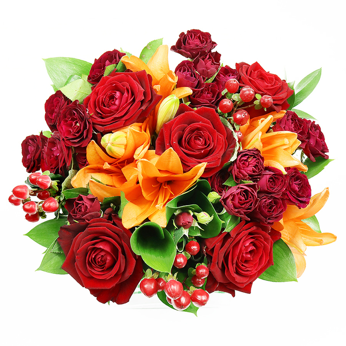 Captured overhead on a spotless white field, this sumptuous circular bouquet feels like a miniature garden of warm tones. At its core, several large, deeply coloured red roses open with layered, velvety petals that suggest traditional romance and a soft, lingering fragrance. Surrounding these are vivid orange Asiatic lilies - some spread wide with their stamens visible, others caught in the graceful arc of an opening bud - adding animated structure and bright trumpet shapes. Tight clusters of smaller burgundy spray roses or miniature garden roses provide concentrated patches of darker hue and a fine, ruffled texture that contrasts with the larger, smoother rose faces. Polished hypericum berries in glossy ruby-red appear as jewel-like accents, their spherical forms reflecting light and introducing a playful, festive temperament. Between the blooms, generous green foliage-broad, glossy ruscus or salal leaves-peeks through, giving the arrangement breathing space and a cool, verdant counterpoint to the warm palette. The composition is dense yet airy, the soft, even illumination revealing tactile differences: the suede softness of rose petals, the velvety sheen of lily tepals, the firm gloss of berries, and the satiny leaves. Designed to convey love, celebration, or seasonal warmth, this bouquet sits equally at home on a Bayswater mantelpiece near Lancaster Gate or as a heartfelt delivery to a nearby flat. It feels handcrafted, thoughtfully balanced, and purposefully chosen to evoke joy and intimate sincerity in equal measure.